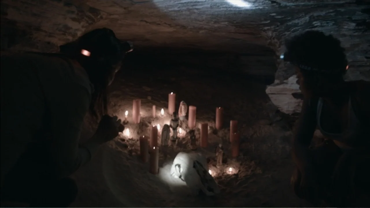 The Grotto (2022) movie still: medium — A dimly lit cave interior features a makeshift shrine with numerous burning candles. Two individuals kneel…
