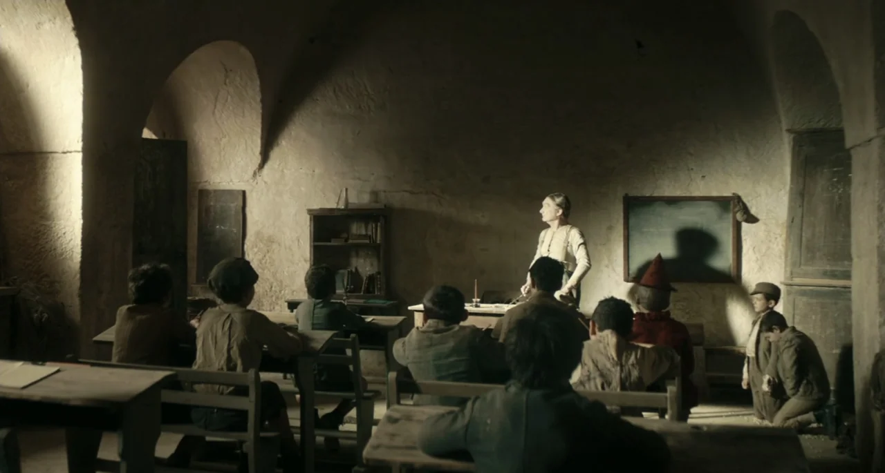 Pinocchio (2022) movie still: wide — A rustic stone-walled classroom features arched ceilings and old wooden school desks. A female teacher stands…
