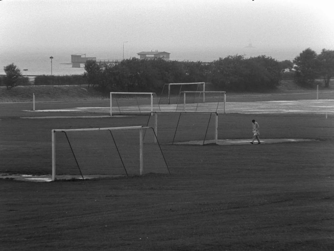 The Baby Carriage (1963) movie still: wide — A large, overcast outdoor soccer field features multiple metal goals on uneven grass. A lone figure…