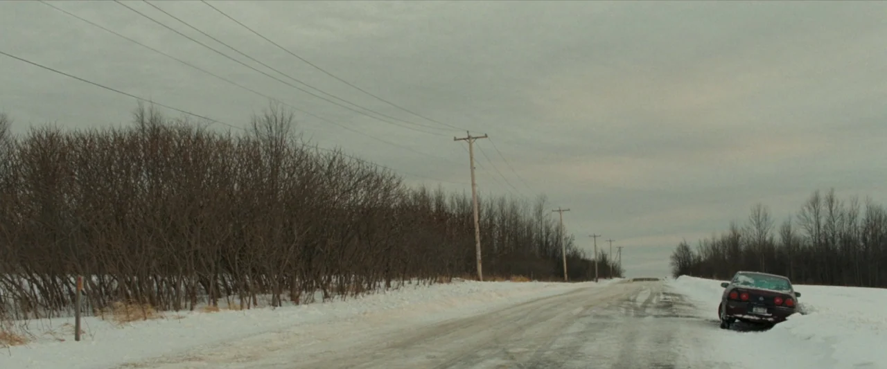 For Ellen (2012) movie still: wide — A desolate snow-covered road stretches past leafless trees under a heavy gray sky. A dark sedan is positioned…