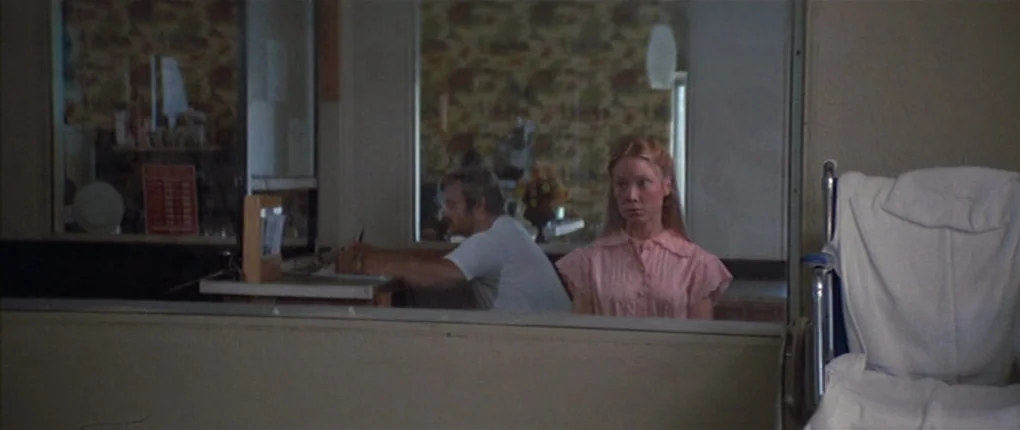 3 Women (1977) movie still: medium — A hospital administrative area featuring a desk, a patient wheelchair, and floral wallpaper. A woman stands…
