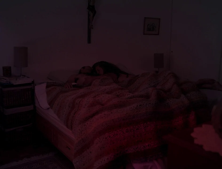 Post Tenebras Lux (2012) movie still: wide — A dark bedroom at night with a crucifix mounted on the wall above the bed. Two people sleep under a…