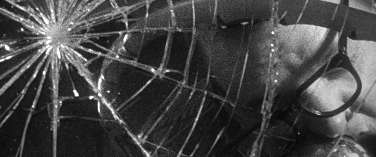 JFK (1991) movie still: closeup — A close view of a shattered glass pane showing sharp radial fracture lines. Reflections distort the dark,…