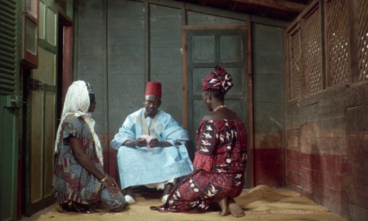 Mandabi (1968) movie still: fullbody — A small room features vertical wooden slat walls and a straw-matted floor. A man in a blue kaftan and red fez…