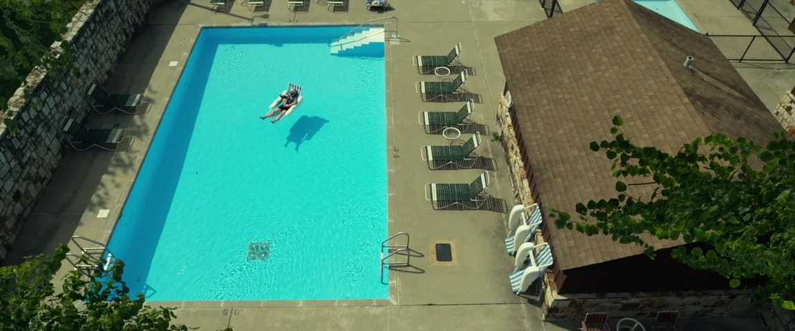 Gone Girl (2014) movie still: wide — An outdoor swimming pool area with green lounge chairs and a small pool house. A lone woman rests on a white…