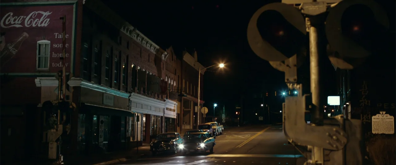 Coming Through the Rye (2016) movie still: wide — A quiet town street at night features red brick storefronts and vintage cars parked. The empty…