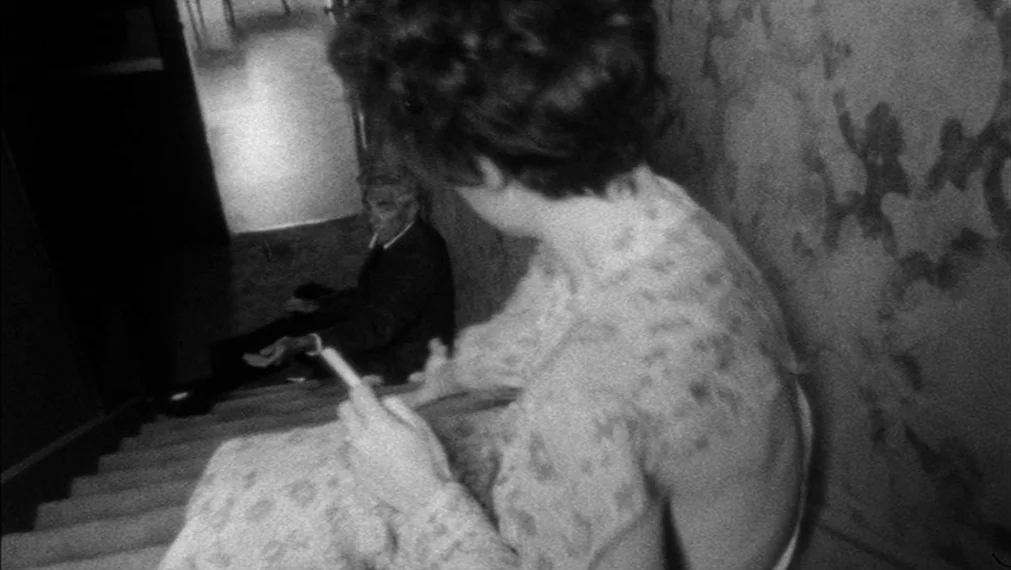 Faces (1968) movie still: medium — A dark, high-contrast interior staircase with patterned wallpaper and carpeted steps. A woman sits on the stairs…