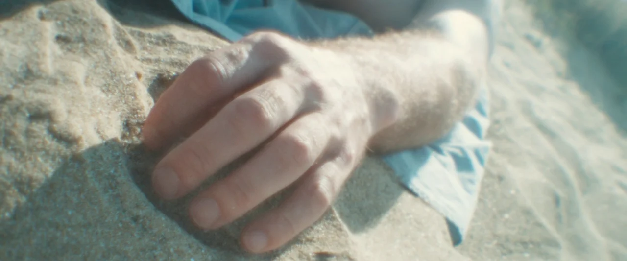 Black Sea (2014) movie still: closeup — A close-up view shows a hand resting in the sand on a bright beach. The person wears a pale blue garment…