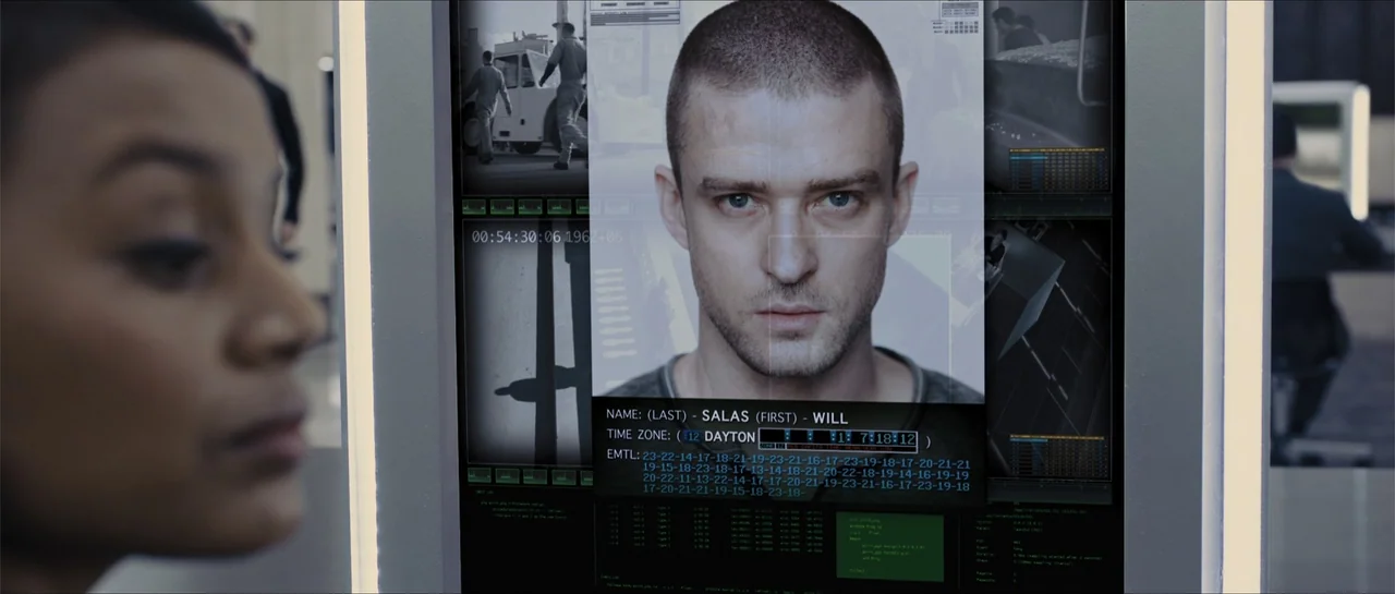 In Time (2011) movie still: medium — A high-tech workspace features a glowing monitor displaying a man's digital identification profile. A woman's…