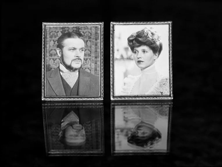 Effi Briest (1974) movie still: closeup — Two rectangular silver-bordered picture frames sit side by side on a reflective black table. The…