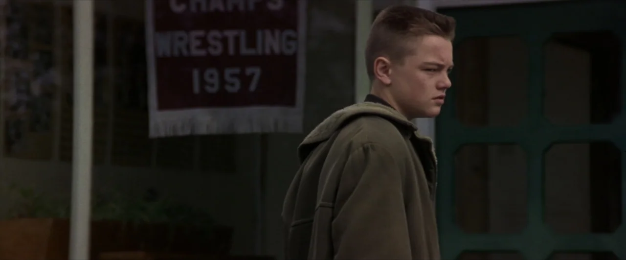 This Boy's Life (1993) movie still: medium — A school entrance features a prominent wrestling poster mounted on a glass door. A teenage boy stands…