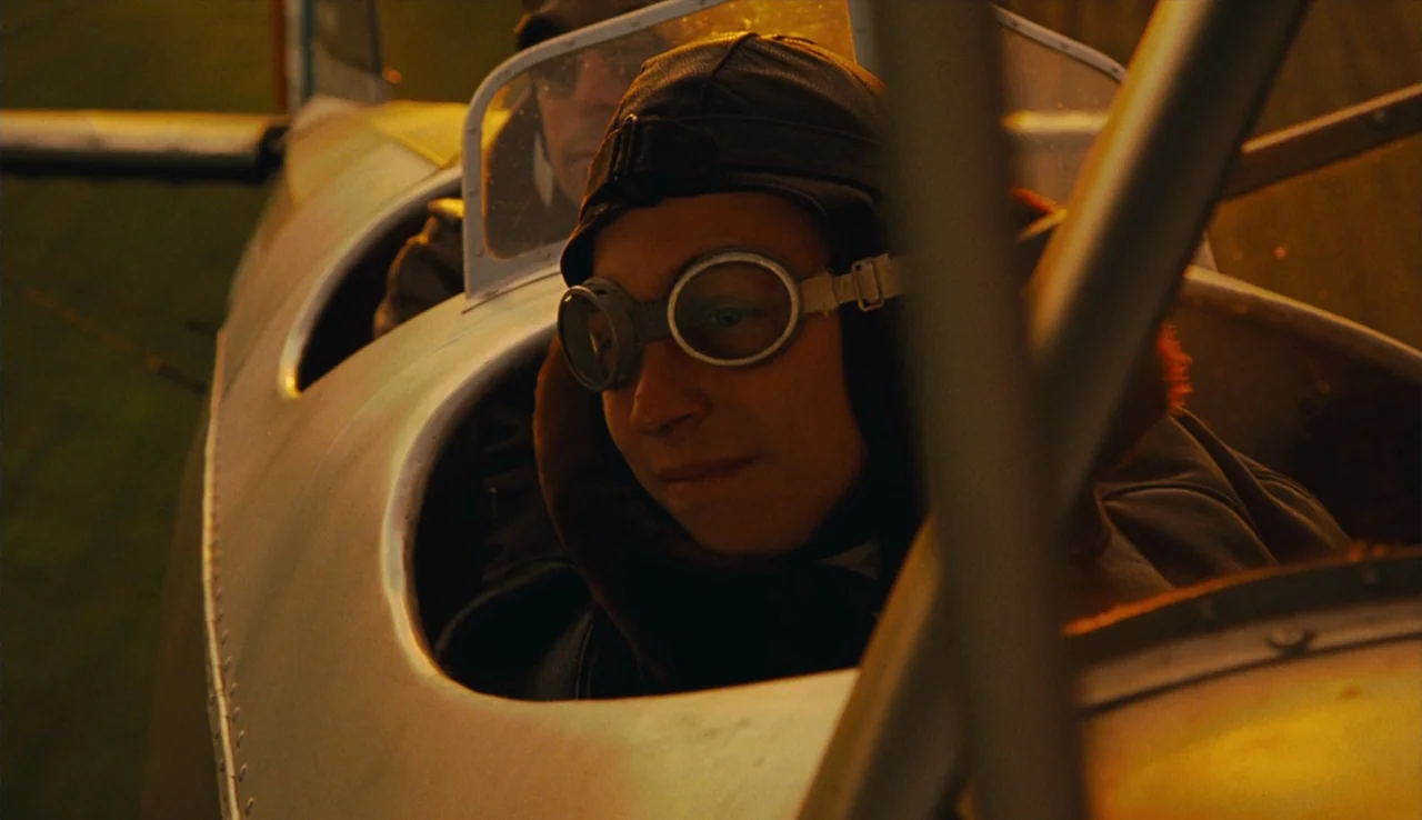 Renoir (2012) movie still: medium — A metal airplane cockpit sits in the open air during a sunset. Two pilots wearing vintage leather headgear and…