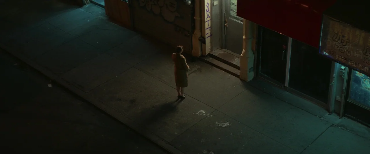 Fair Play (2023) movie still: wide — A nighttime city sidewalk beside a storefront with graffitied walls and a red awning. A woman in a tan coat…