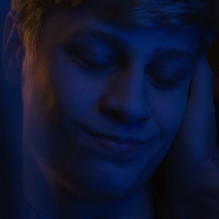 Mommy (2014) movie still: closeup — A close-up view of a young man in a darkened space. The person rests their head in their hand while eyes are…