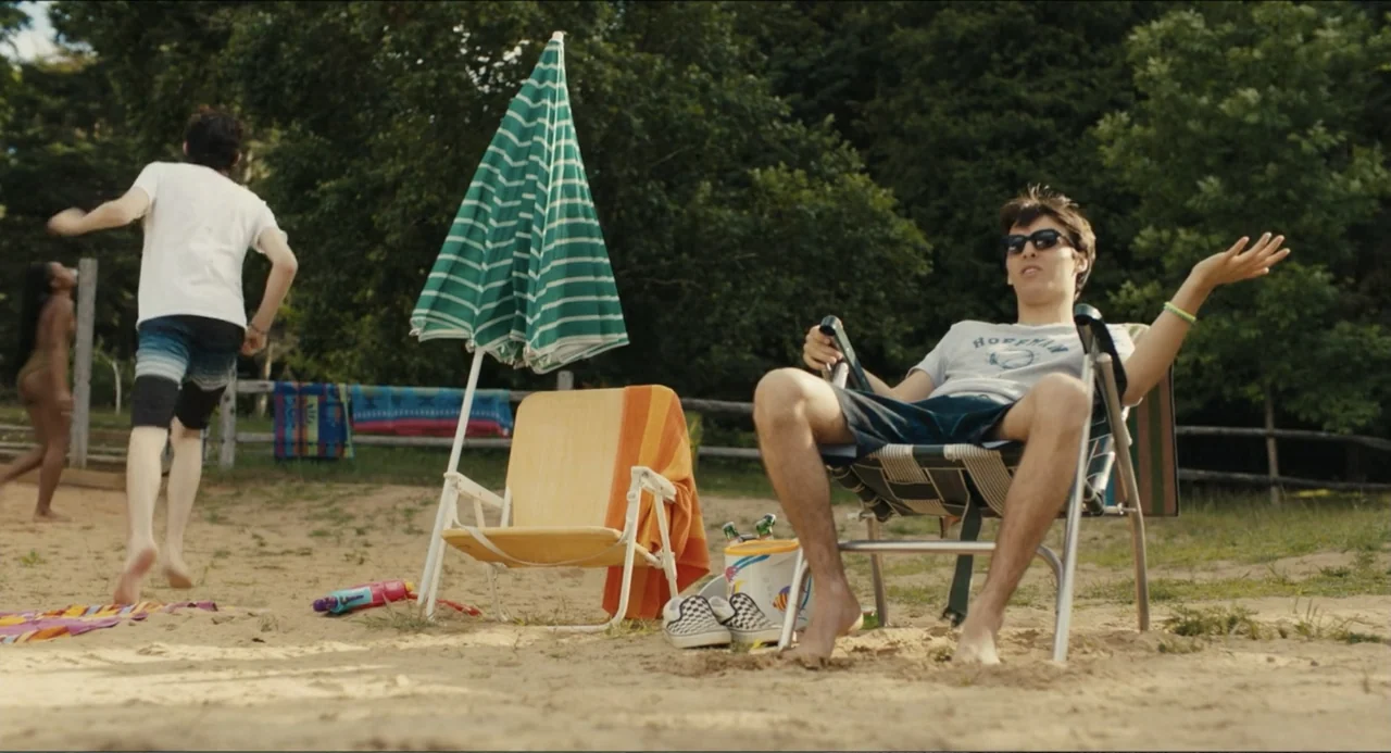 Hell of a Summer (2025) movie still: medium — A sandy beach setting features a wooden fence and dense trees in the background. A young man sits in a…