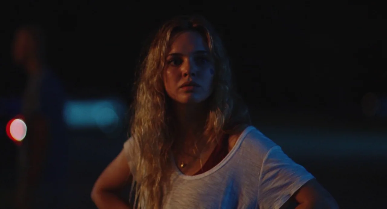 The Giant (2019) movie still: medium — A dark outdoor parking lot at night with blurred colored lights in the background. A woman in a white t-shirt…