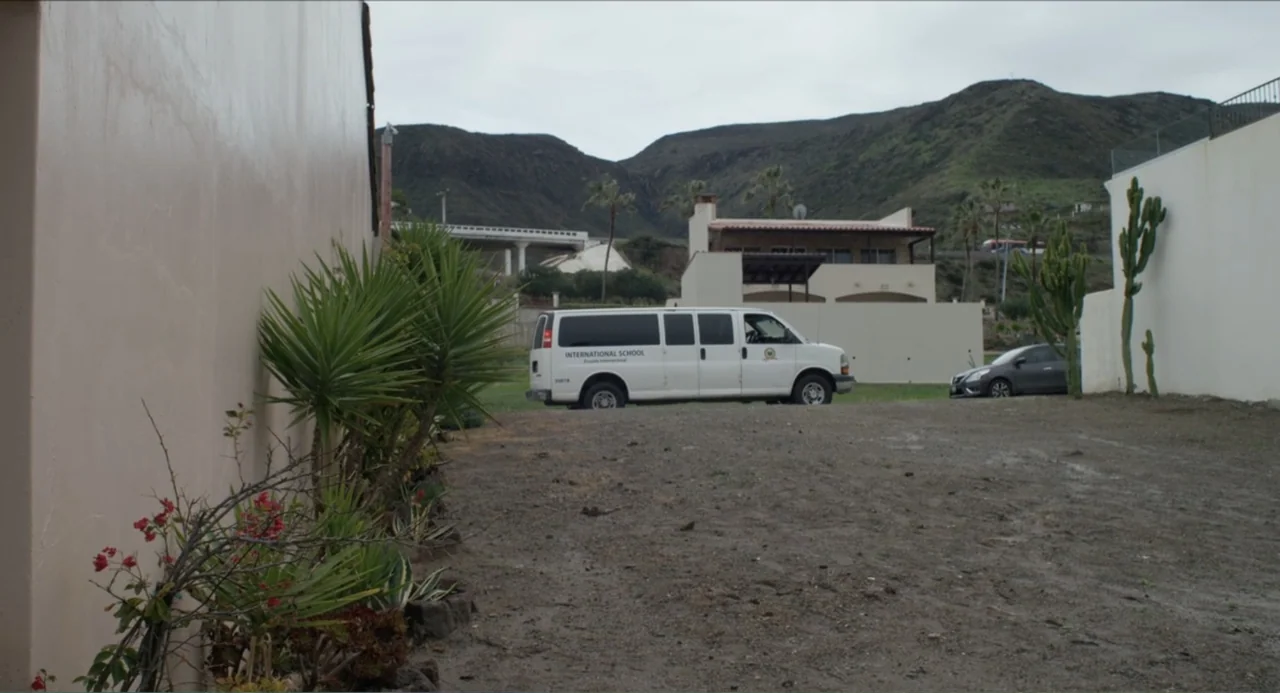 Instantes Capturados (2022) movie still: wide — An overcast exterior dirt lot sits before a white wall and distant hills. A white passenger van…