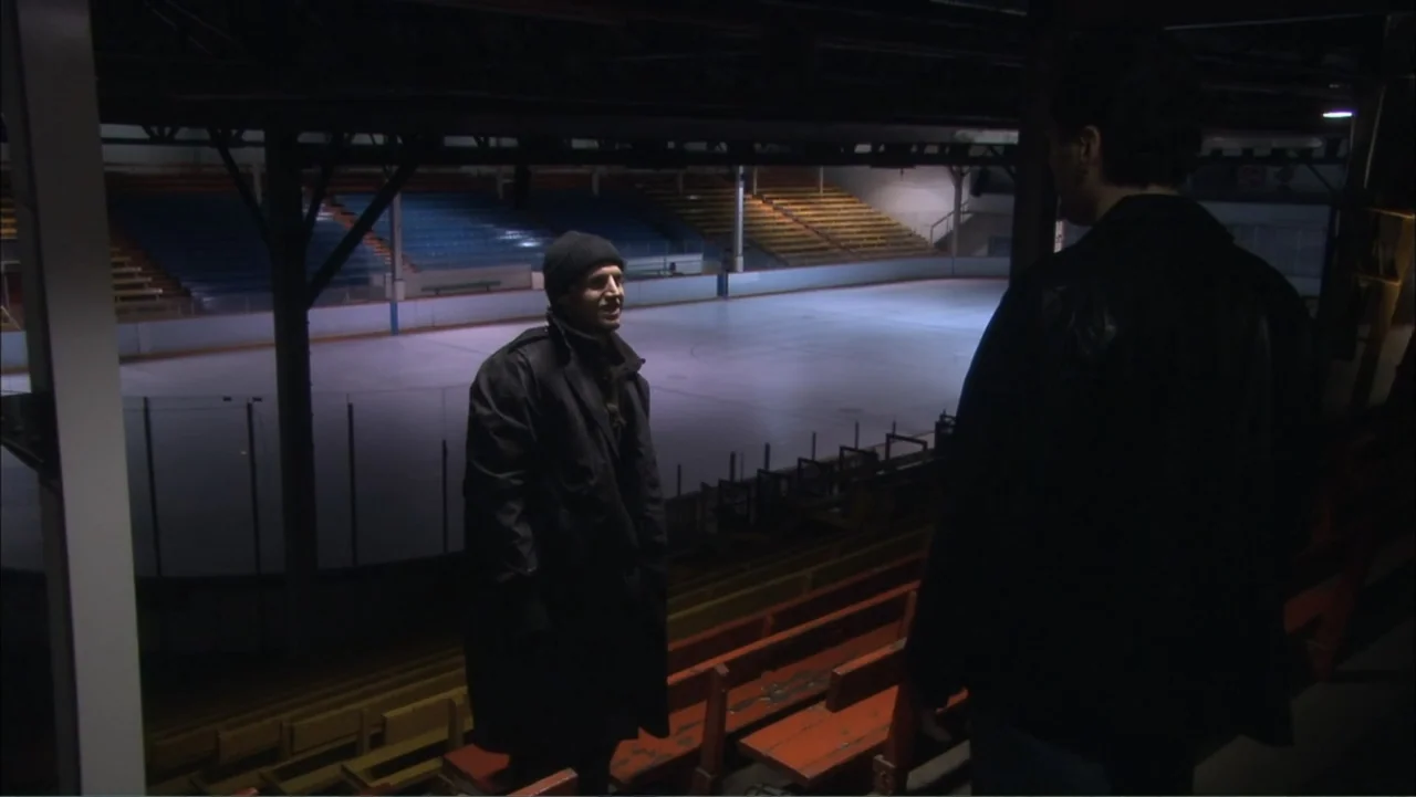 O código oculto (2025) movie still: fullbody — An empty indoor ice hockey arena with tiered wooden bleachers and dark metal rafters. Two men stand…