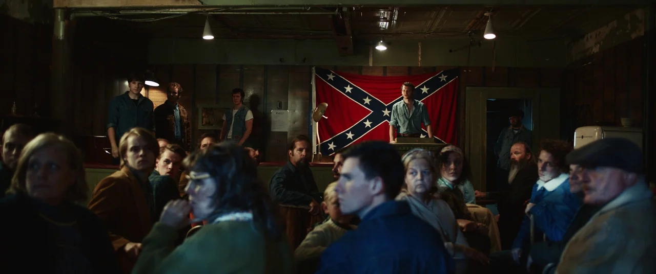 Judas and the Black Messiah (2021) movie still: wide — A dimly lit wooden meeting hall features a large confederate flag draped on the wall. A young…