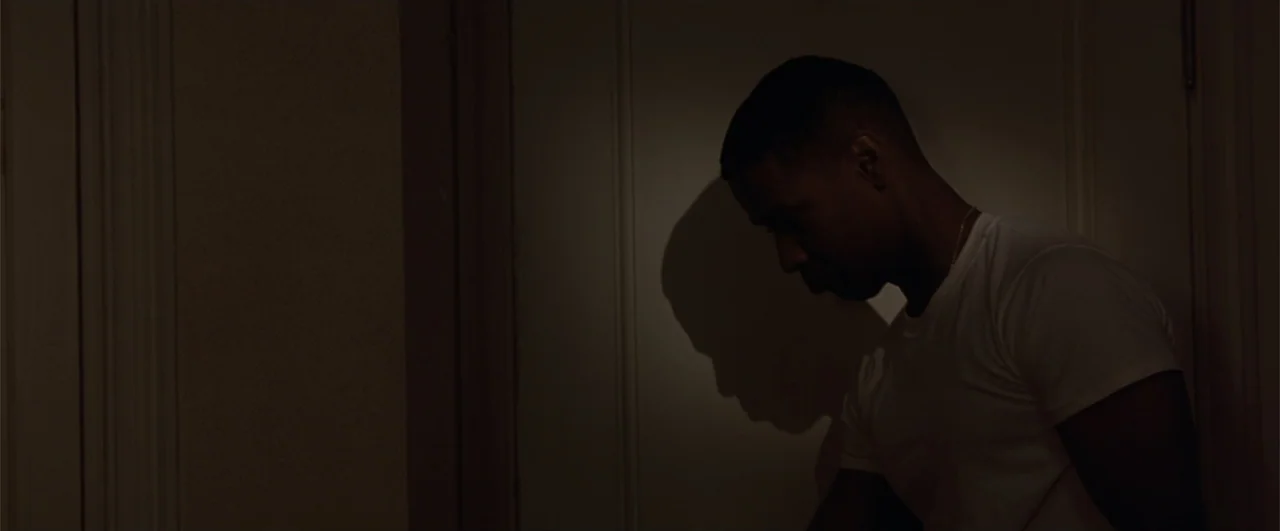Fallen (2004) movie still: medium — A dimly lit interior hallway features a plain wall and a wooden door frame. A man in a white t-shirt stands near…