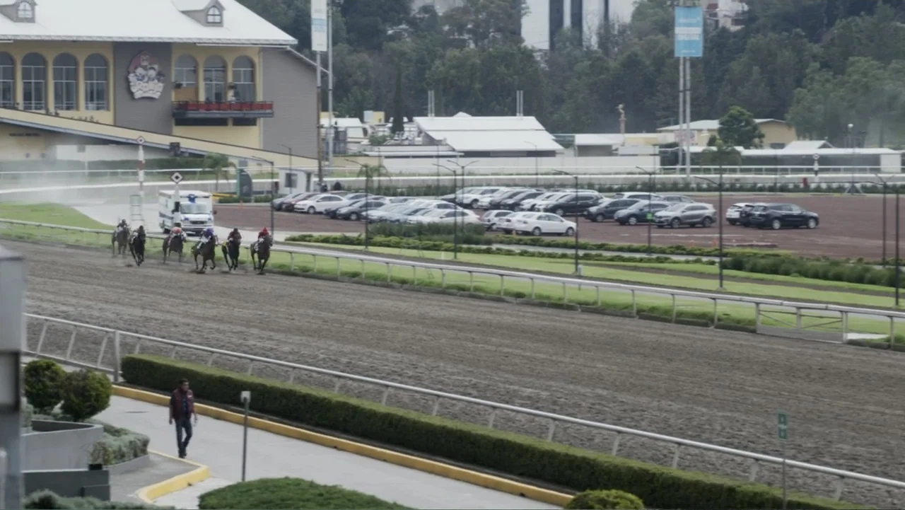 Lecciones para canallas (2022) movie still: wide — A wide view of a dirt horse racing track with a pedestrian walkway. Several jockeys on horses…