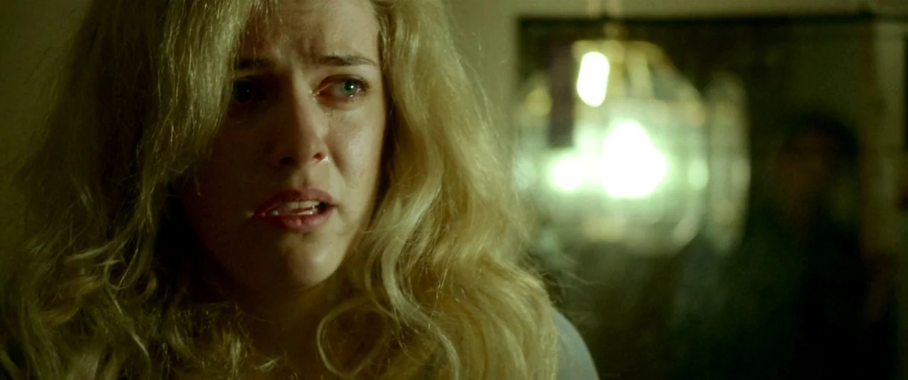 The House That Jack Built (2018) movie still: closeup — A dimly lit room contains a glowing practical light fixture in the background. A blonde…