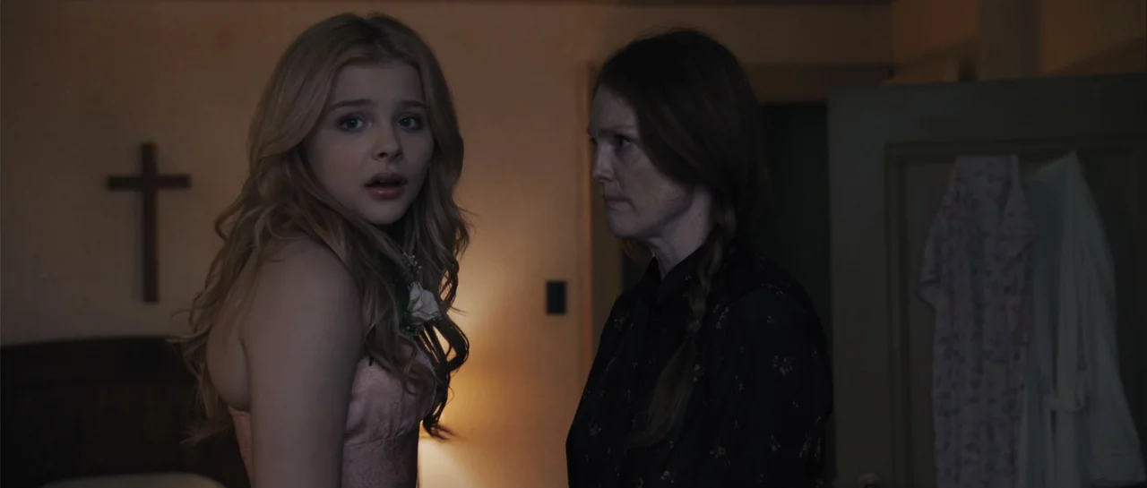 Carrie (2013) movie still: medium — An interior domestic space featuring a wall cross and a hanging robe. Two women face each other in a dimly lit…