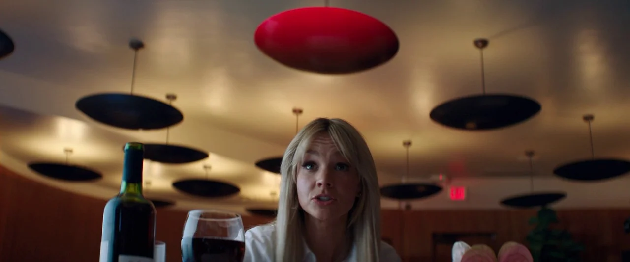 Promising Young Woman (2020) movie still: medium — A woman sits at a table in a modern space with numerous dark hanging lamps. She is talking while…