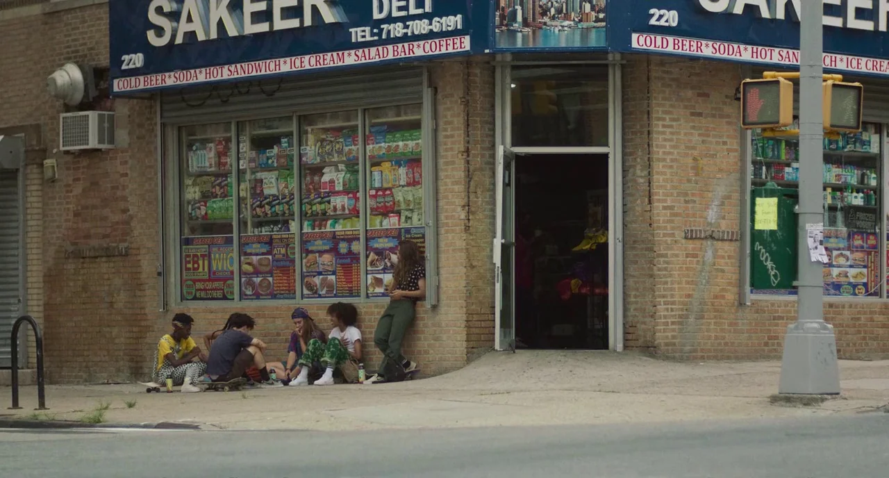 Skate Kitchen (2018) movie still: wide — A corner storefront with a blue sign featuring advertisements and a phone number. Five young people sit on…