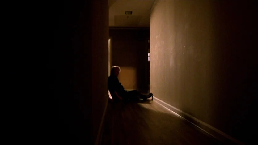 Love Liza (2002) movie still: wide — A long, dark domestic hallway with walls receding into deep shadows. An older man sits on the floor near the…