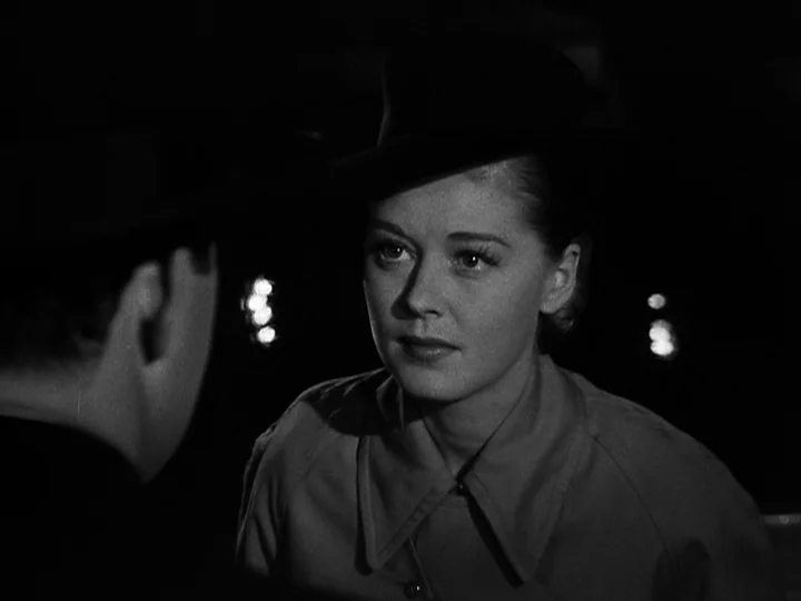 Nightmare Alley (1947) movie still: medium — A dark, blurred outdoor nighttime environment with distant out-of-focus city lights. A woman in a hat…