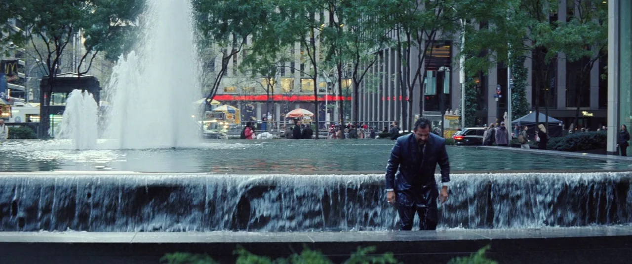 Uncut Gems (2019) movie still: wide — An urban plaza in New York City features a modern stone fountain with water. A man in a fully drenched dark…