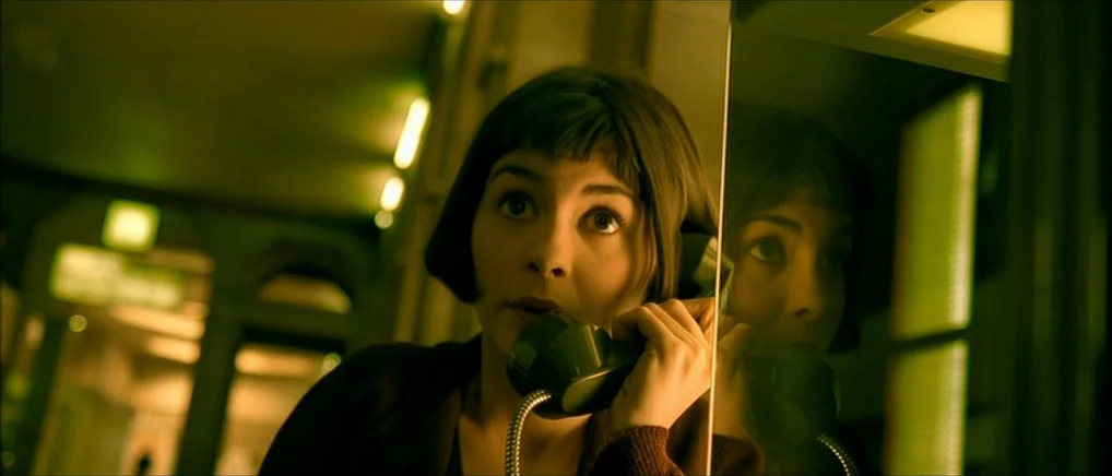 Amelie (2001) movie still: closeup — A woman stands in a small, enclosed public space with a mirror on the wall. She is talking on a landline…