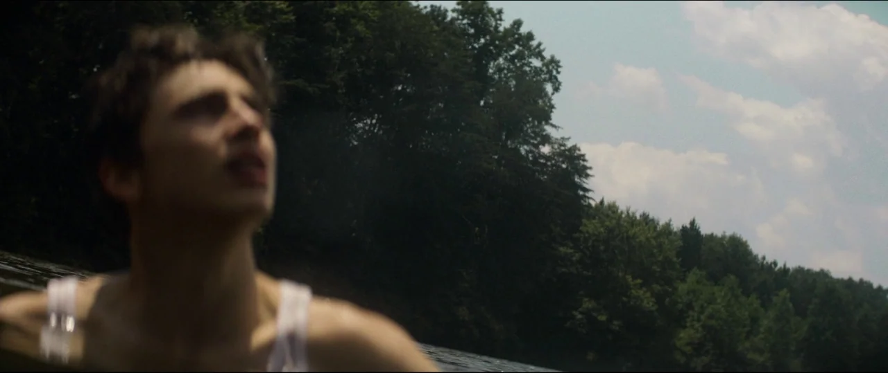 One & Two (2015) movie still: closeup — A body of water is framed by a line of dense green trees. A young man wearing a white tank top looks up…