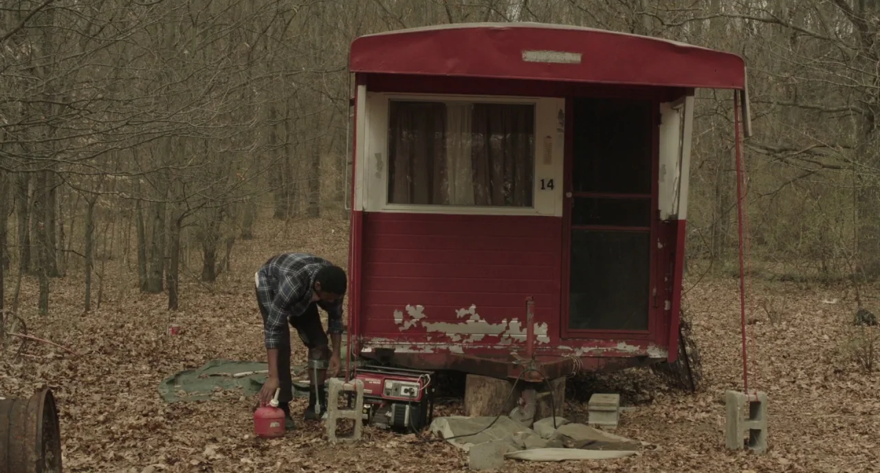 The Alchemist Cookbook (2016) movie still: wide — A small weathered red metal trailer sits in a clearing of a deciduous forest. A man crouches…