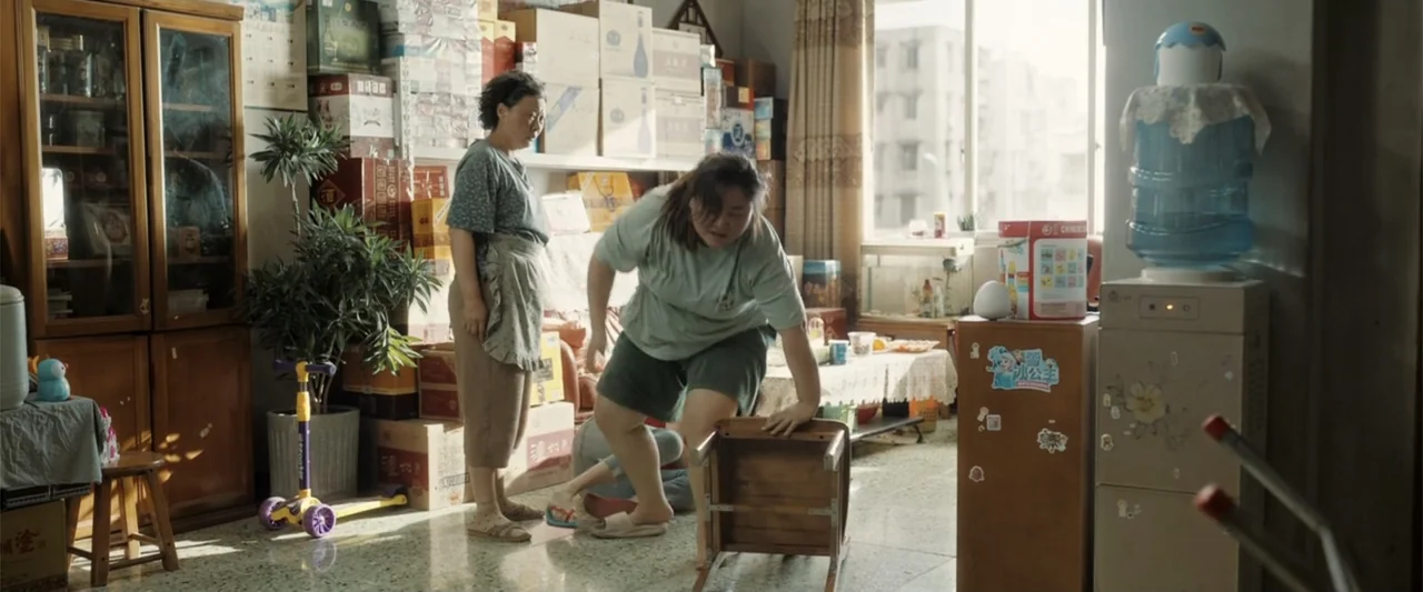 Yolo (2024) movie still: medium — A cluttered apartment living room features stacks of cardboard boxes and a water cooler. One woman stands while…