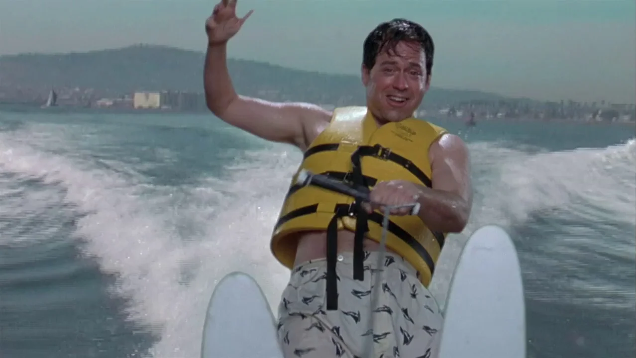 Auto Focus movie still: medium — A man water skis across an open body of water with distant coastal hills visible. He wears a yellow life vest and…
