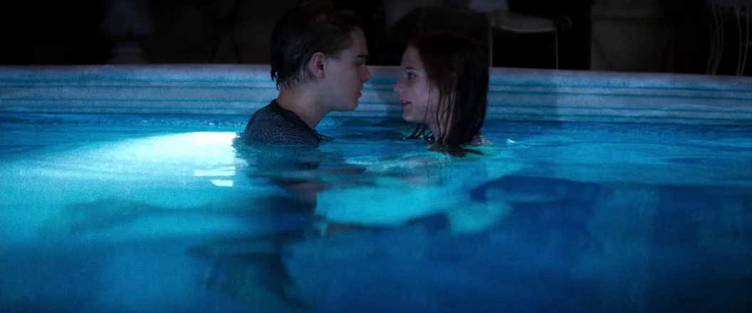Romeo + Juliet (1996) movie still: medium — A man and woman stand in a illuminated pool at night. The water reflects the underwater lighting.