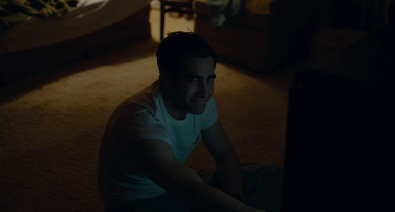 Wildlife (2018) movie still: medium — A dark, sparsely furnished room with carpeted floors is illuminated primarily by screen light. A man wearing a…