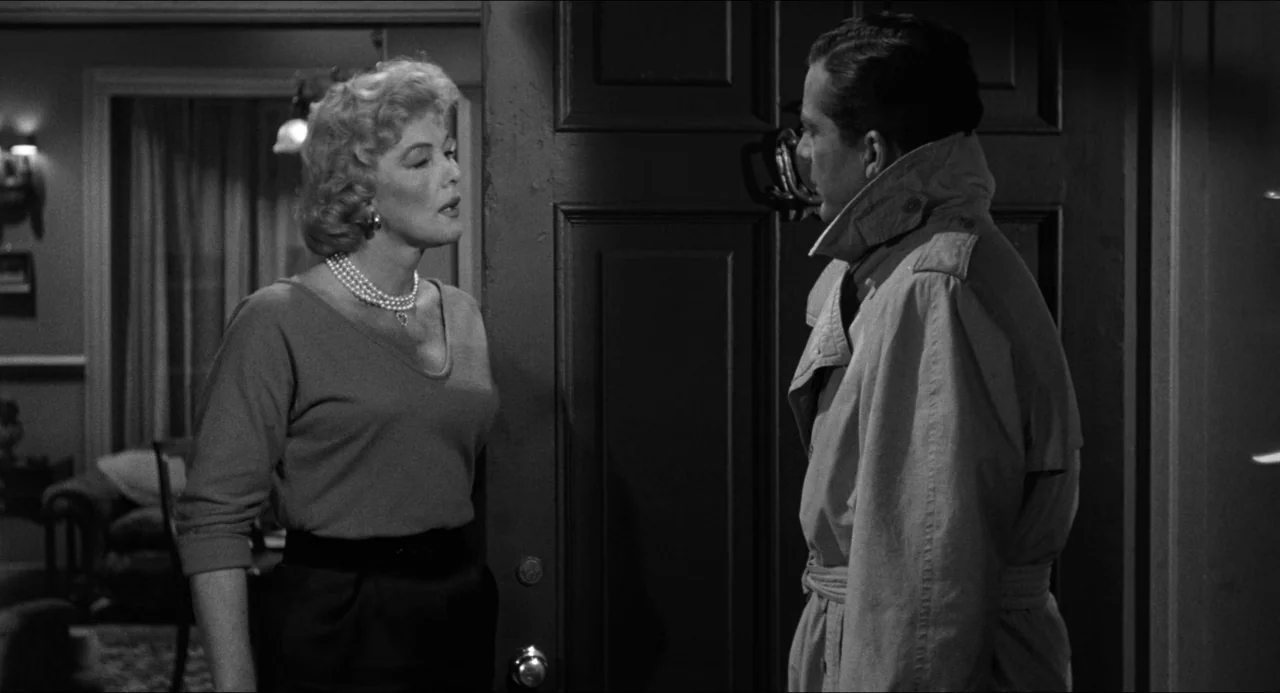 The Fearmakers (1958) movie still: medium — An interior entryway features a dark wooden door separating the two subjects. A woman in a sweater faces…