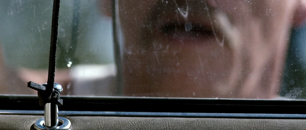 Cop Car (2015) movie still: closeup — A close-up view through a car window with a manual door lock handle visible. A blurred face presses against…