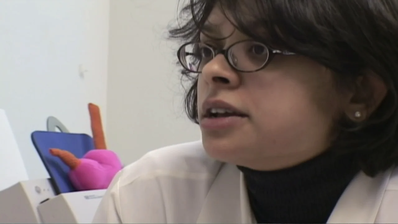 Super Size Me (2004) movie still: closeup — A sterile office environment features a white wall and a white desk surface. A woman in a lab coat looks…