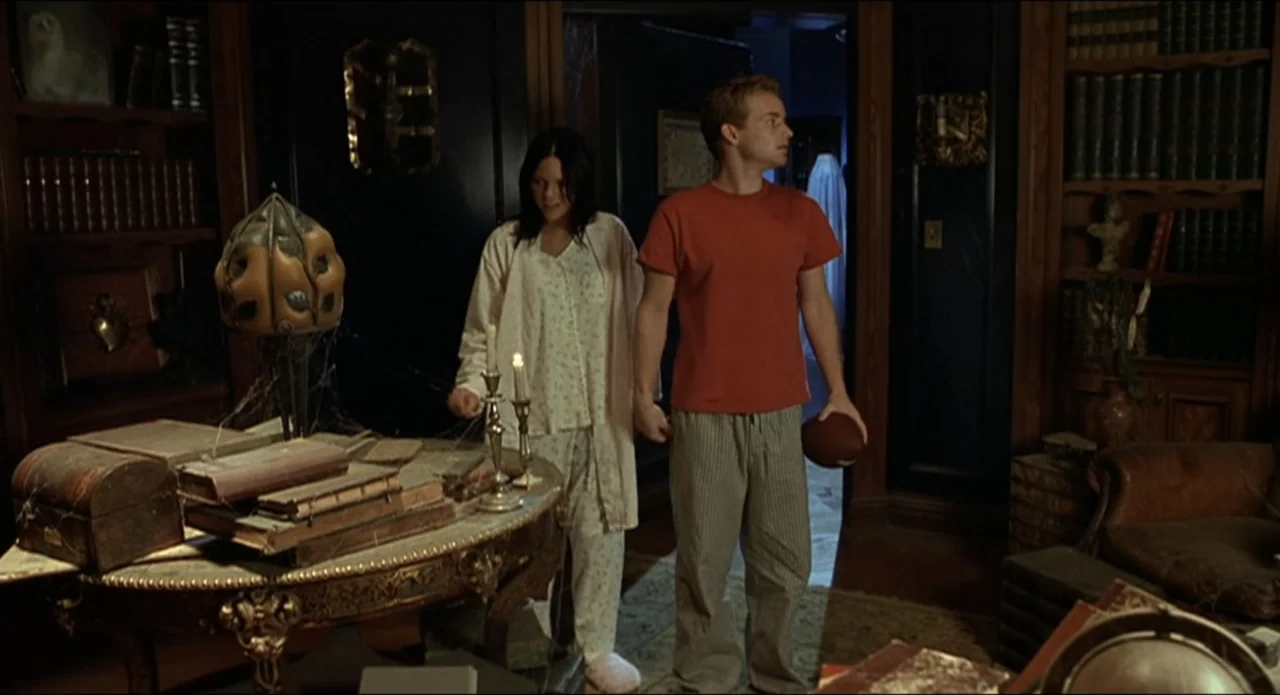 Scary Movie 2 (2001) movie still: medium — A dark library features dark wood bookshelves filled with books and a cluttered central table. A man and…
