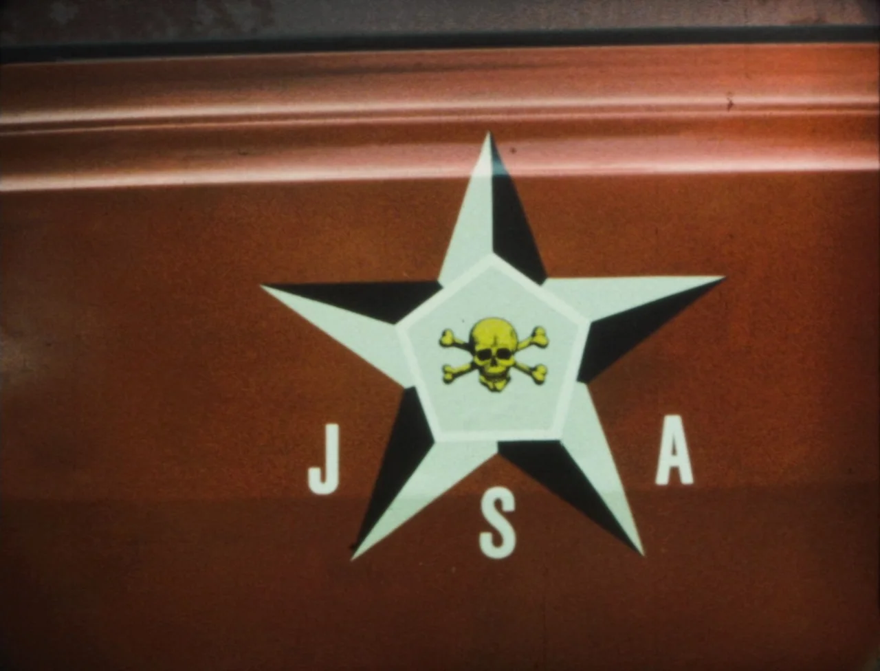 Nekromantik (1988) movie still: closeup — A close-up view of a matte rust-colored metal surface with painted graphic markings. The decal features a…