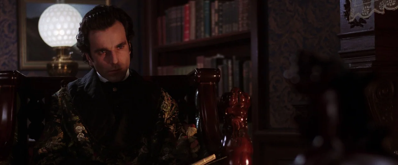 The Age of Innocence (1993) movie still: medium — A dimly lit room with dark wooden bookshelves and ornate wallpaper background details. A man in a…