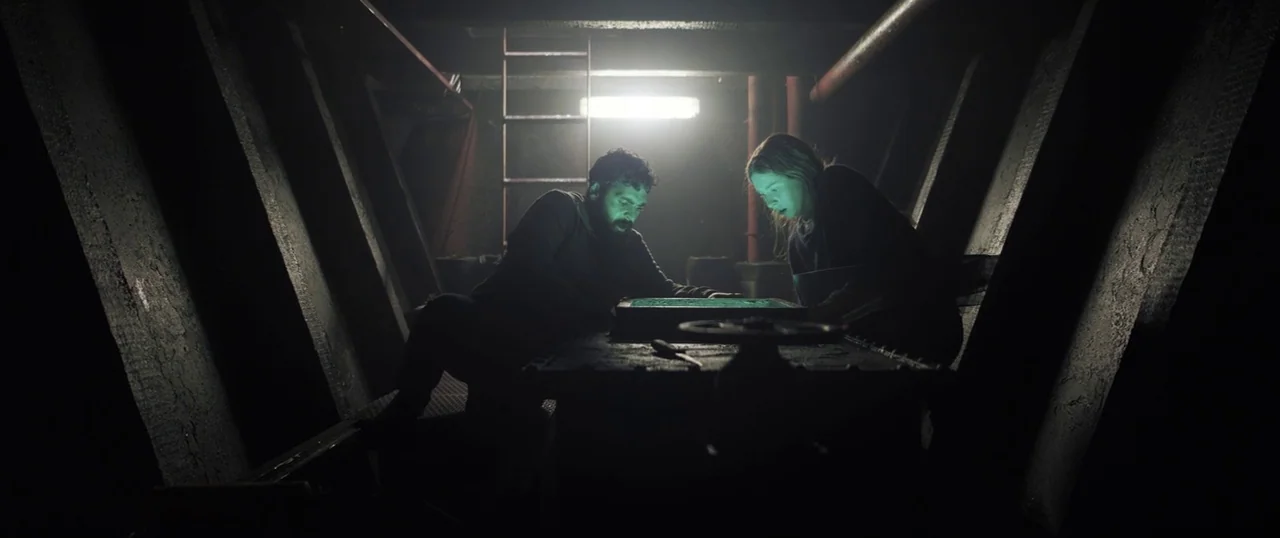 Sea Fever (2019) movie still: medium — A dimly lit metallic industrial compartment features diagonal support beams and a ladder. A man and a woman…