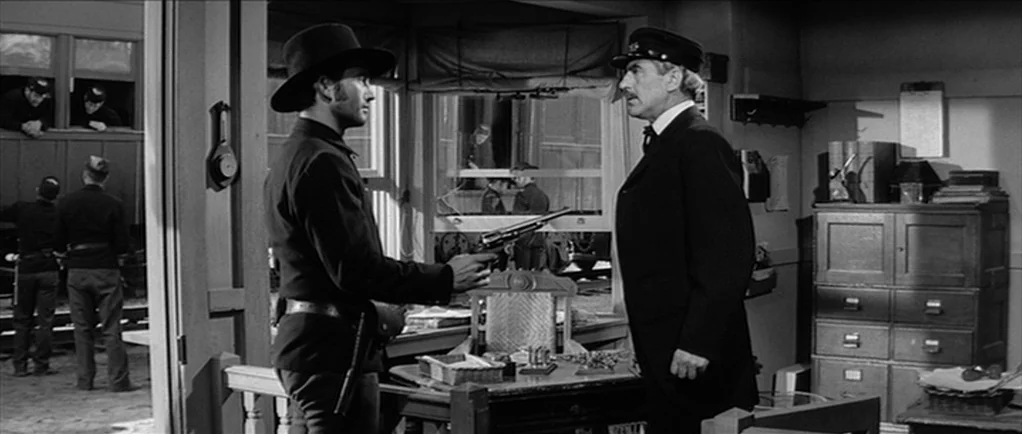 Love Me Tender movie still: medium — A wooden railway station office with a large window and heavy filing cabinets. A man in a cowboy hat points a…
