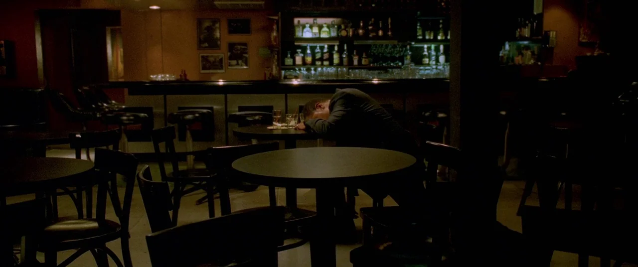 Collateral (2004) movie still: wide — An dimly lit establishment features circular tables and dark chairs facing a back bar. A man rests his head on…