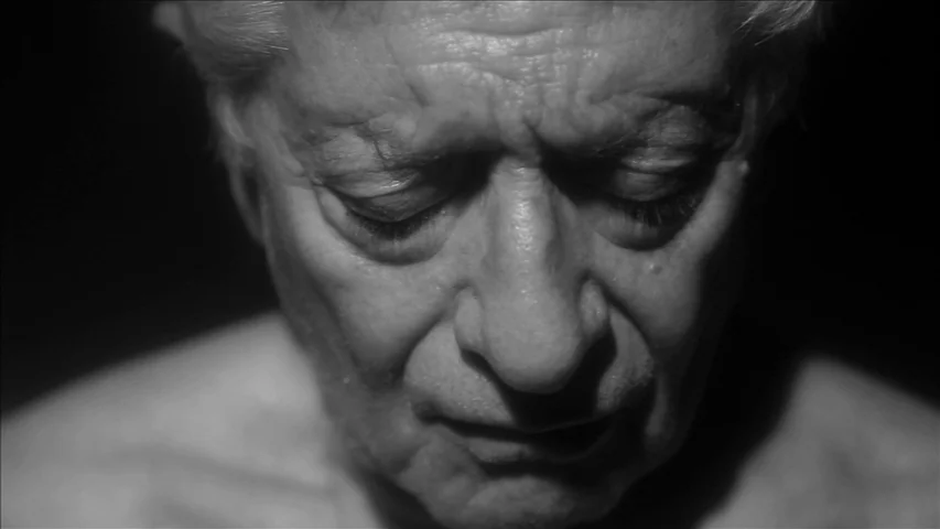 Keyhole (2011) movie still: closeup — An elderly man is framed in a dark, indistinct interior setting with minimal light. He keeps his head angled…