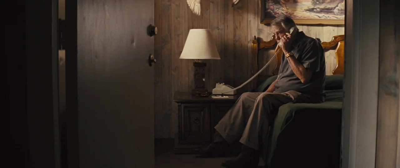Mud (2012) movie still: medium — A small room features wood-paneled walls, an ornate lamp, and a landscape painting hanging above. An older man sits…